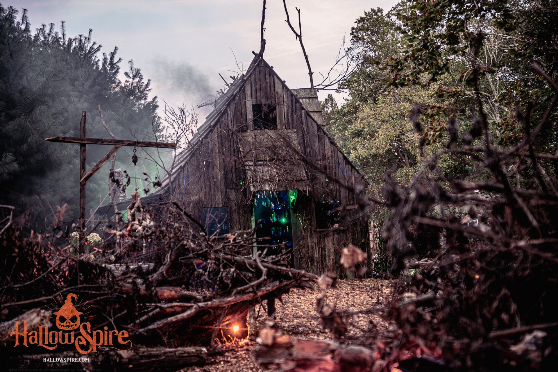 A crooked wooden cottage at Hallowspire surrounded by twisted branches, fog, and orange and green lighting.